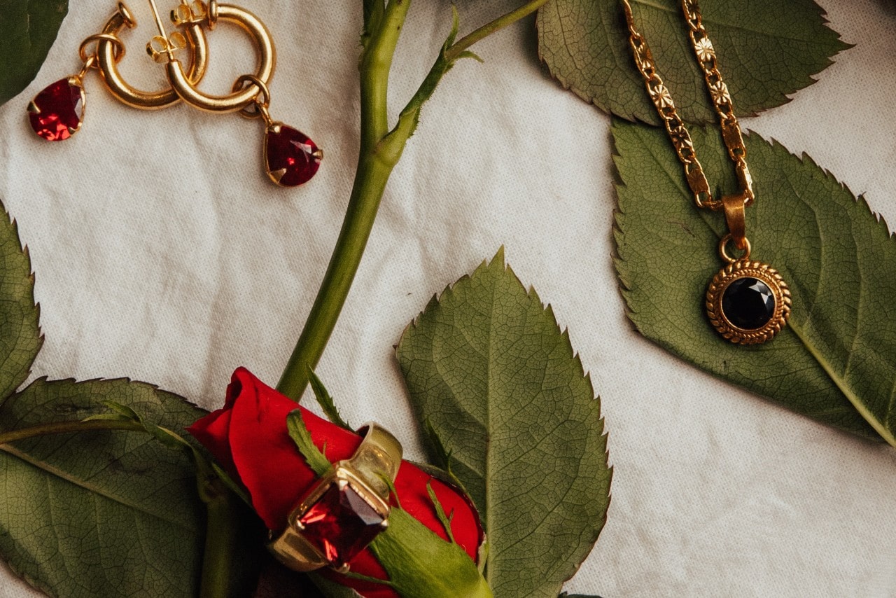 Ruby and yellow gold drop earrings, gemstone fashion ring, and pendant displayed on a single red rose and leaves with a white cloth background