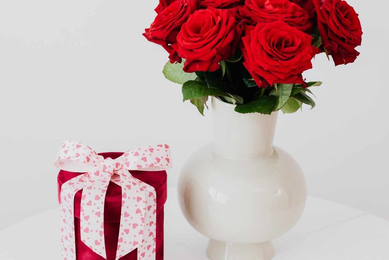 A bouquet of red roses in a white vase next to a velvet cylinder box with a white and pink heart bow on top