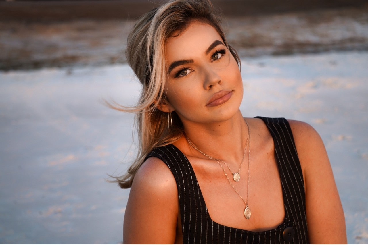 A woman wearing two coin necklaces sits on a white sand beach