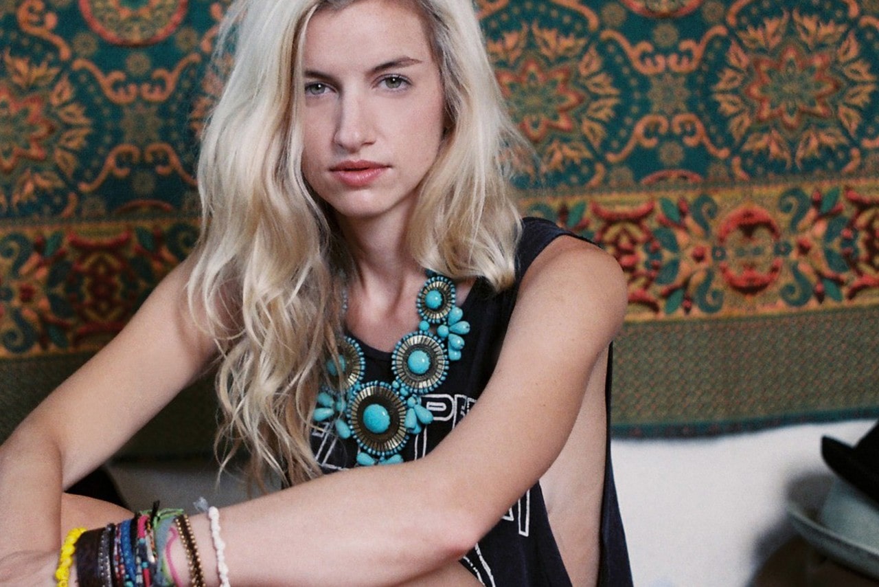 A woman wearing an elaborate turquoise necklace sits by a tapestry