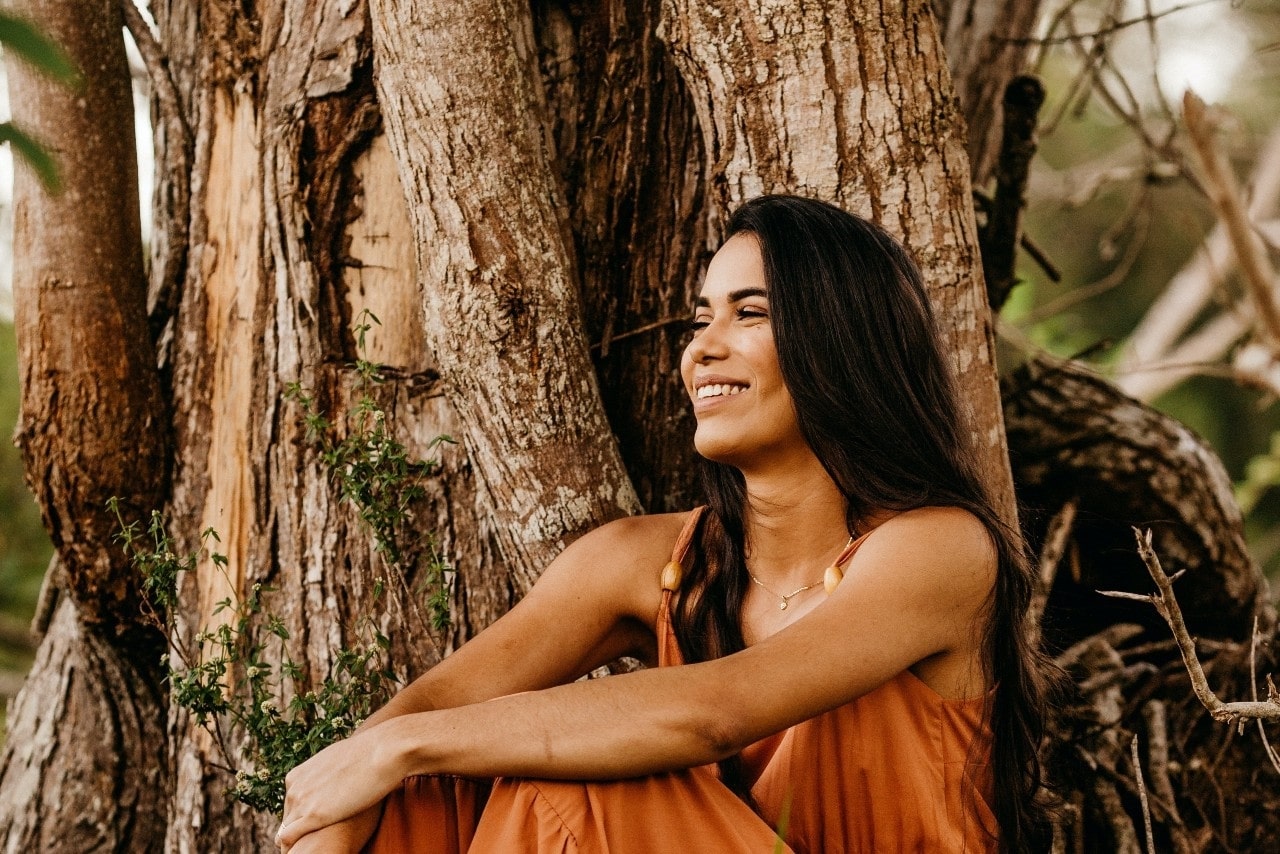 lady smiling, sitting outside against a tree, and wearing a fashionable necklace