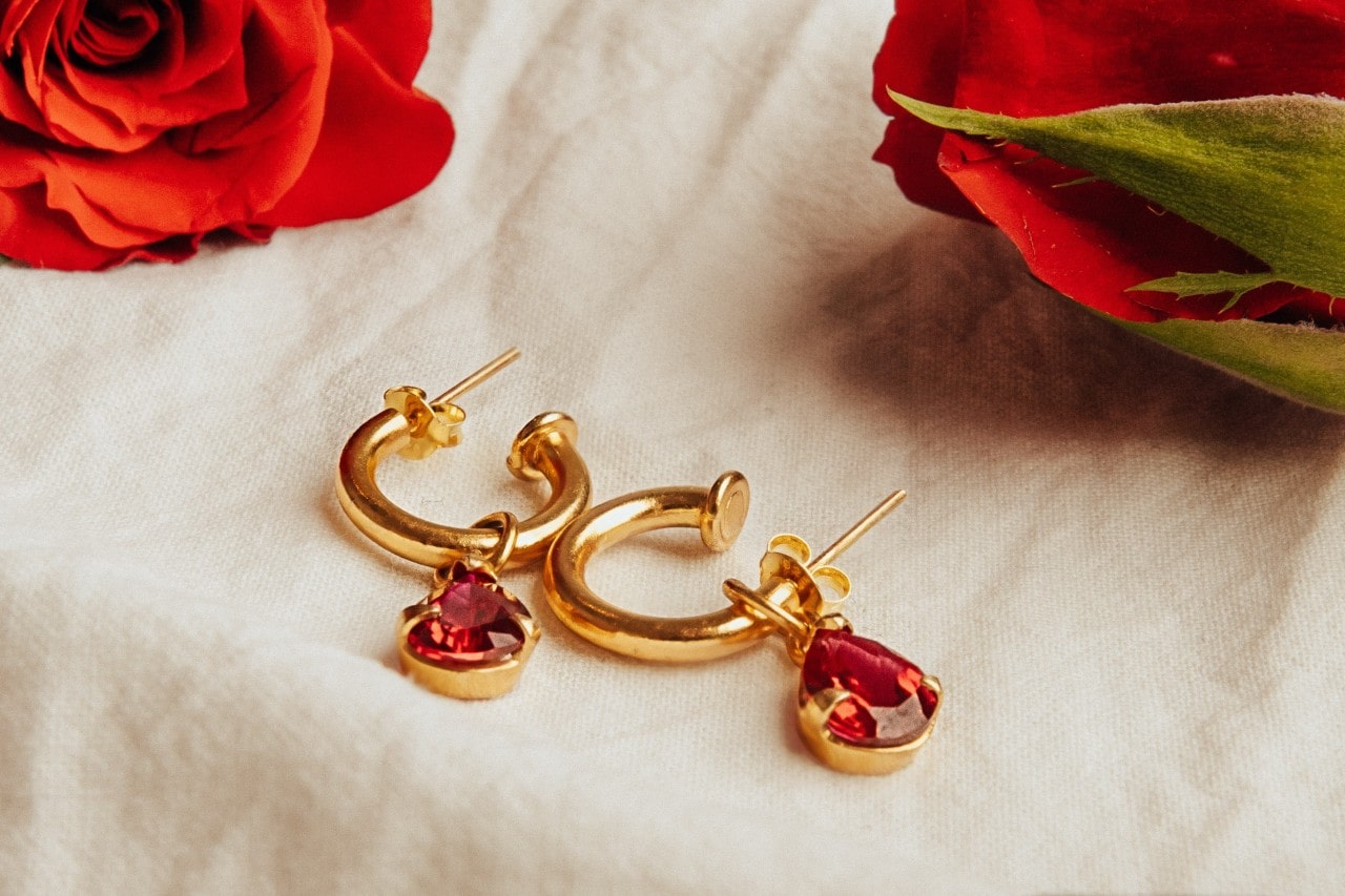 a pair of yellow gold huggie/drop earrings featuring red gemstones lying next to roses