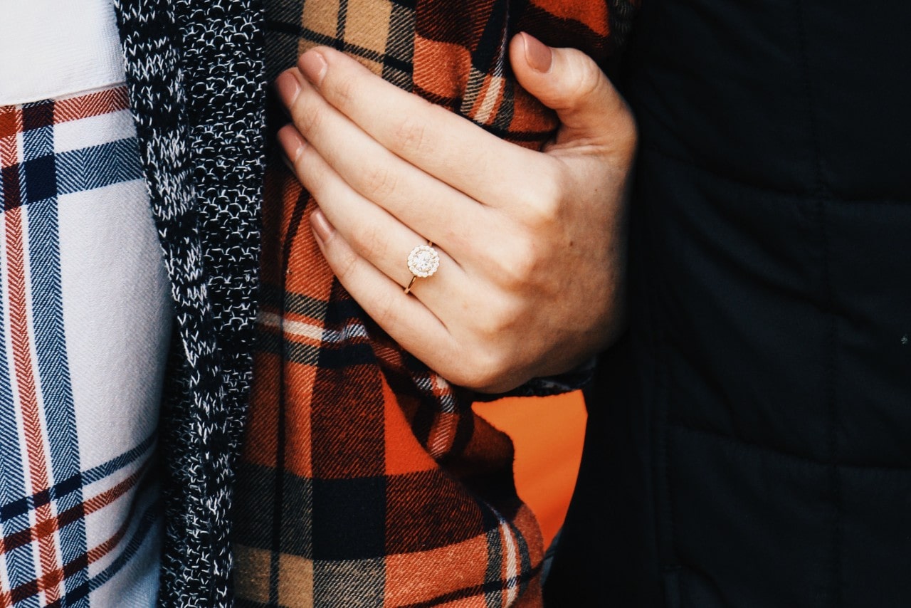 A close-up of a woman’s hand in an engagement ring holding her partner’s arm.