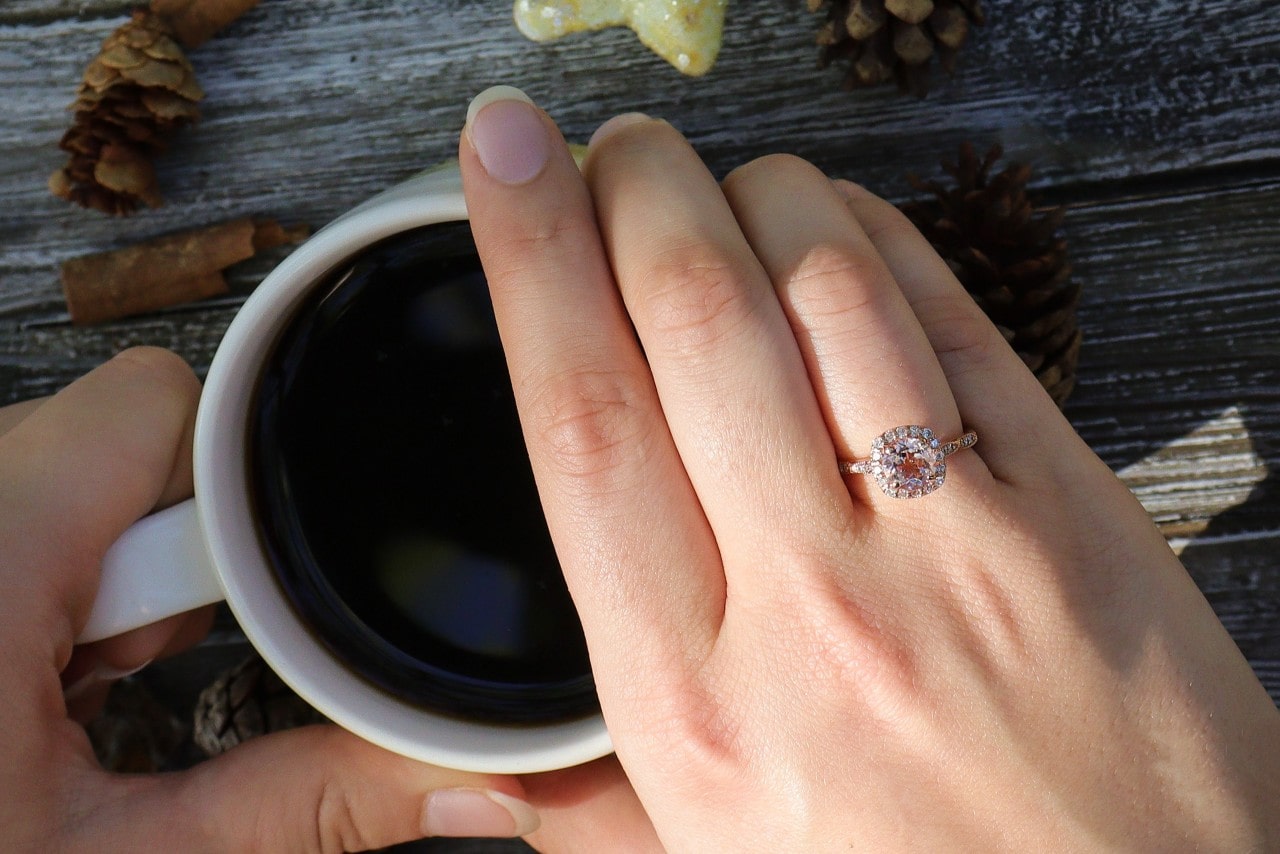 A close-up of a woman’s hand adorned in an elegant engagement ring as she drinks coffee outside in fall.