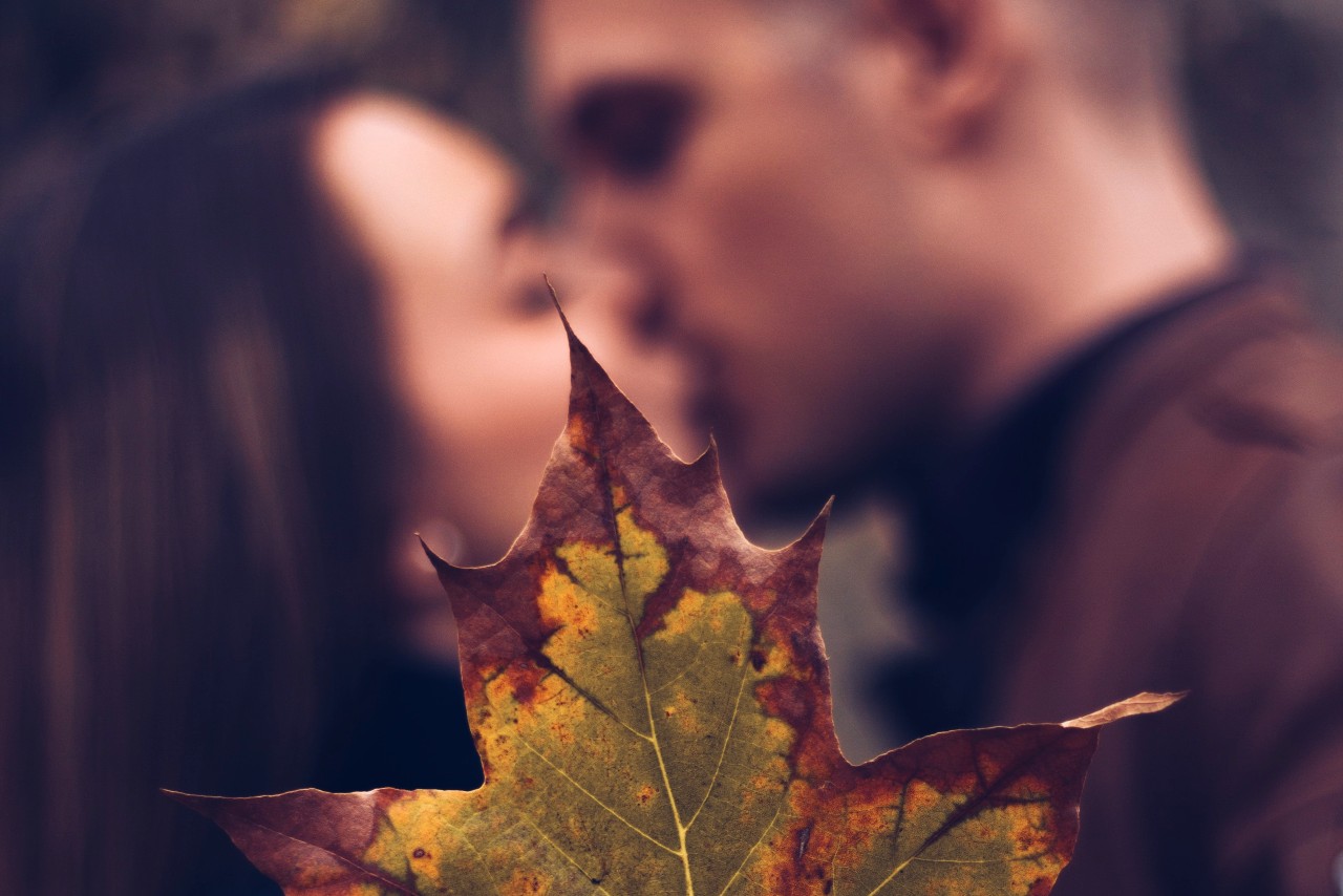 A close-up of a young couple kissing, with an autumn leaf in the foreground.