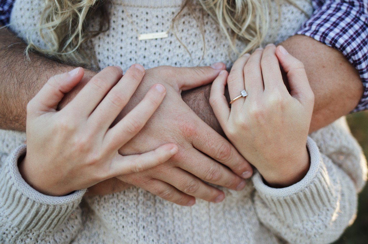 A close-up of a man holding his partner from behind, with emphasis on her necklace and engagement ring.