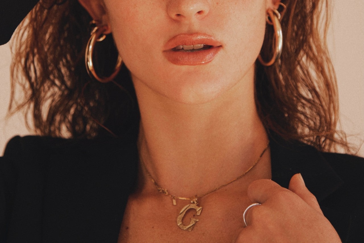 A woman wearing a black blazer, a gold monogram “G” necklace, and gold hoop earrings.