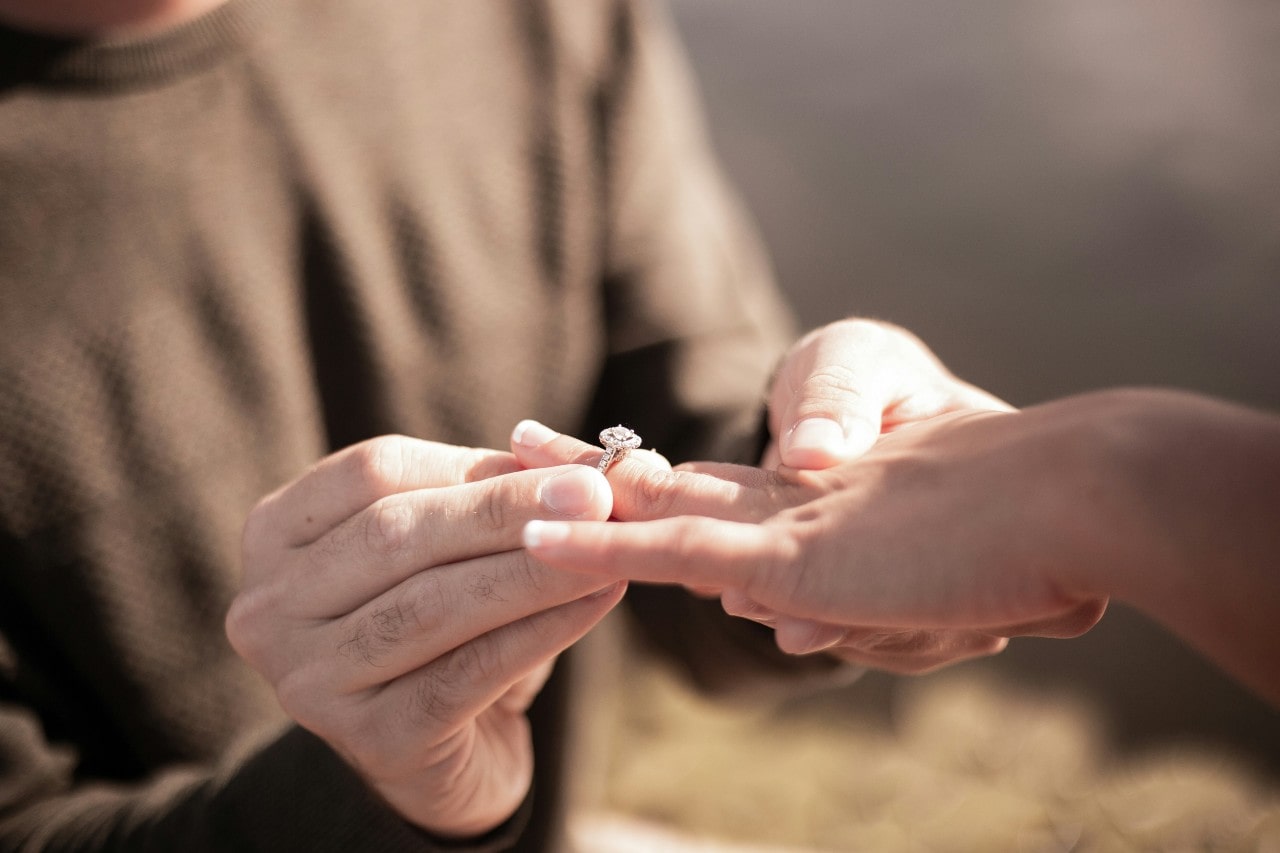 A man in brown weather puts a cushion cut diamond engagement ring on a woman’s finger in the outdoor sunny setting A man in brown weather puts a cushion cut diamond engagement ring on a woman’s finger in the outdoor sunny setting