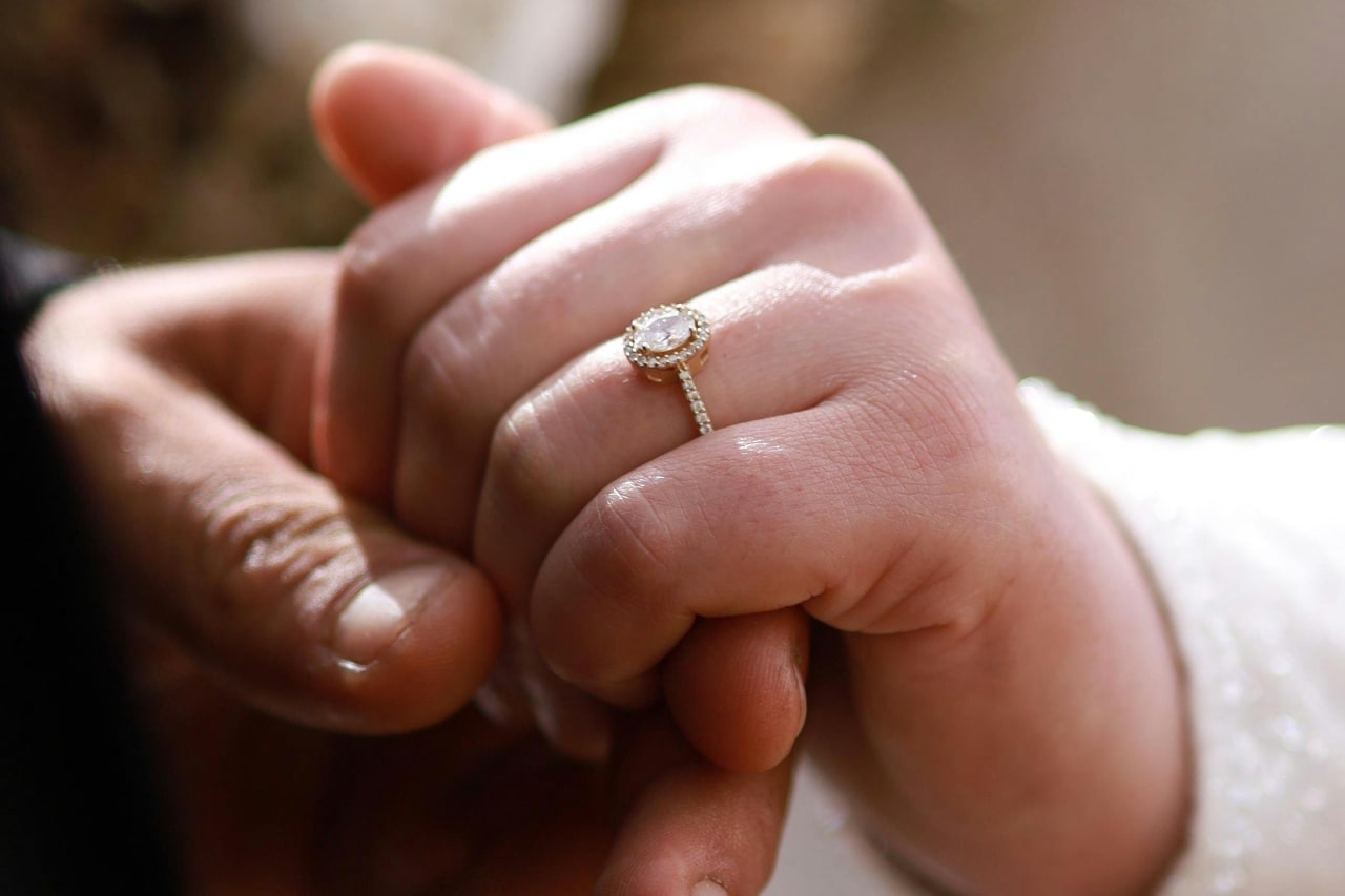 A close-up of a couple holding hands, the bride's hand wearing a white gold engagement ring featuring an oval halo setting. A close-up of a couple holding hands, the bride's hand wearing a white gold engagement ring featuring an oval halo setting.