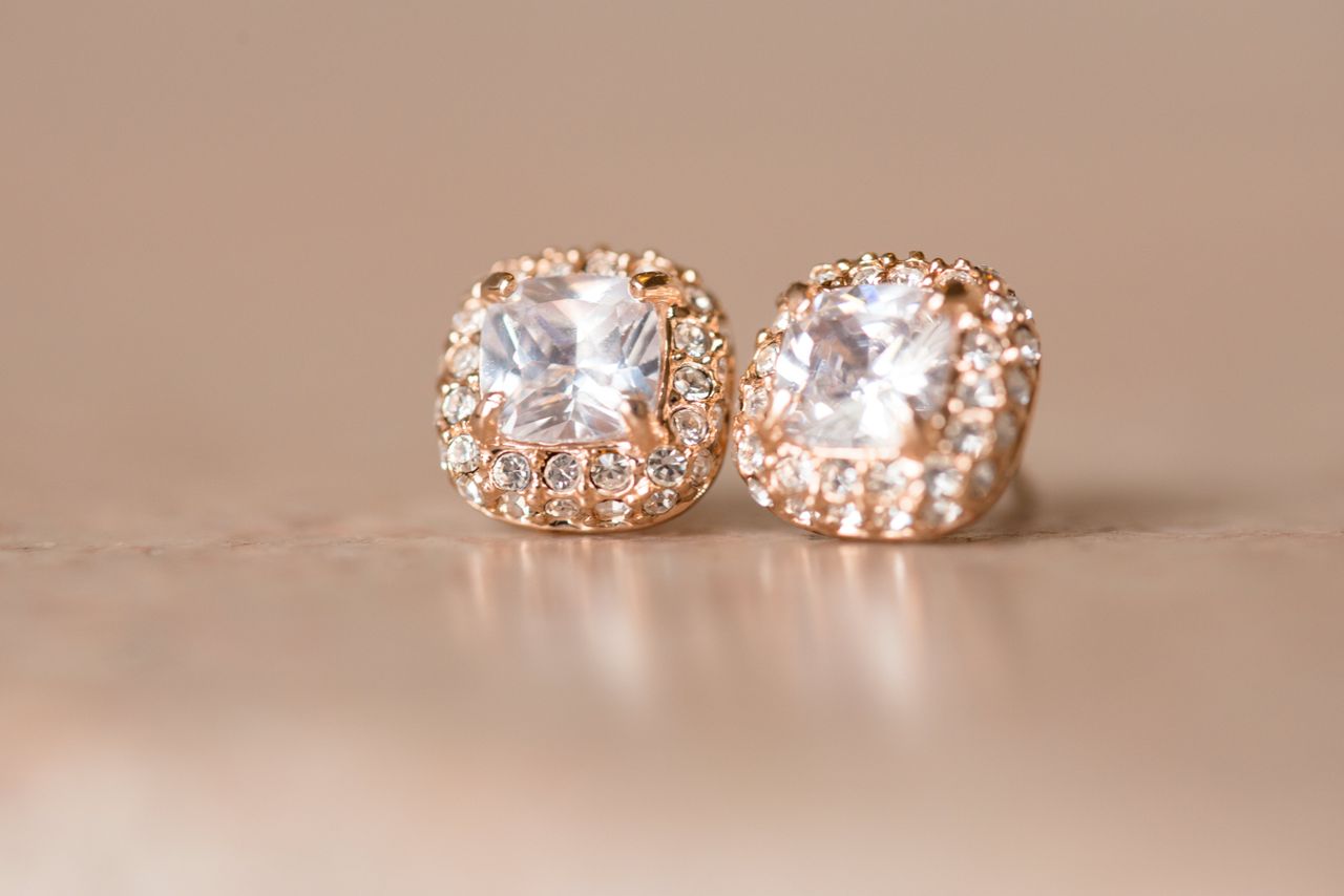 A pair of rose gold earrings adorned with cushion-cut gemstones surrounded by sparkling pavé-set diamonds on a beige background.