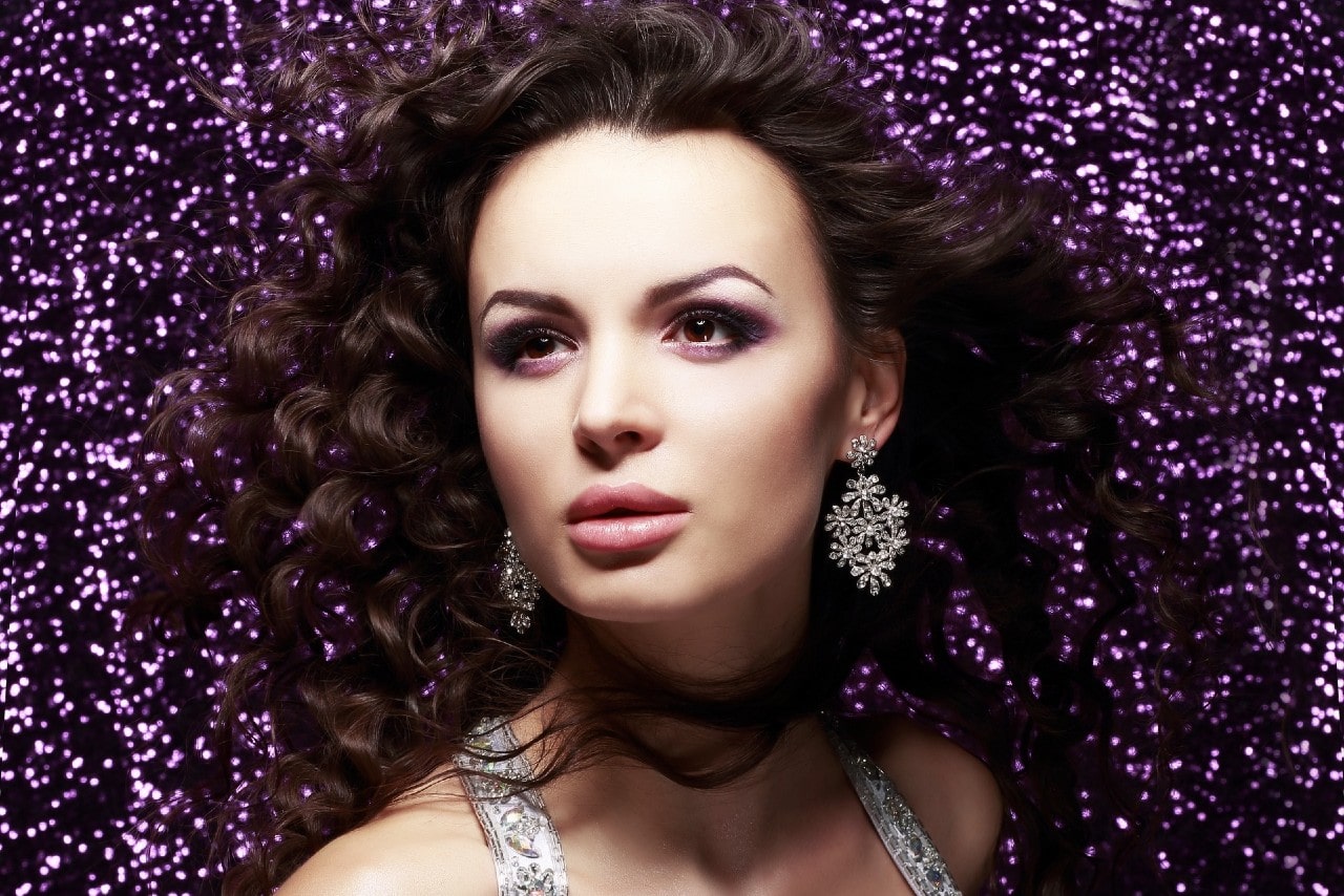 A portrait of a woman with voluminous curly hair, wearing ornate, sparkling chandelier earrings against a shimmering purple background.