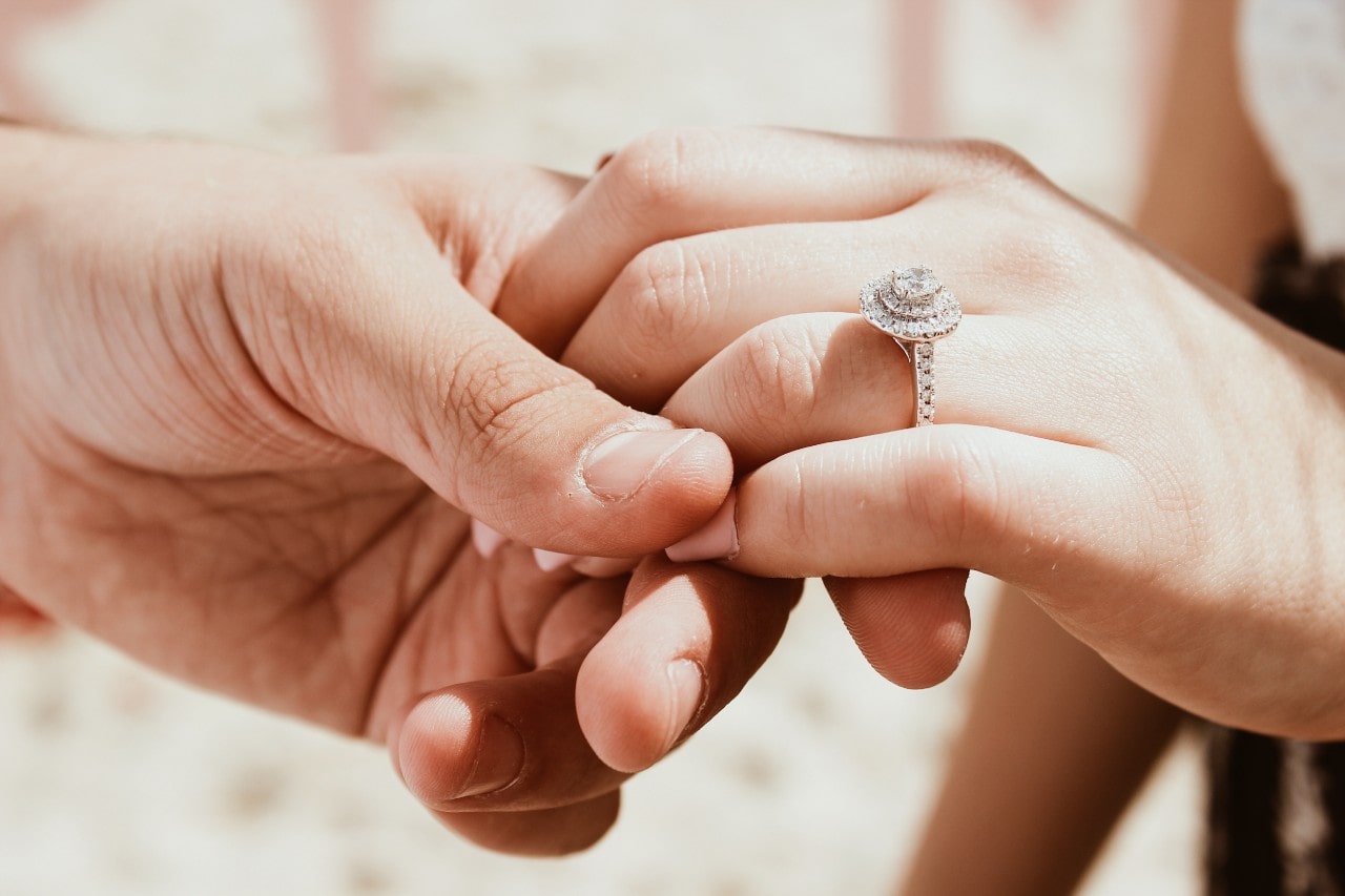 A close-up of two hands clasped together, with a sparkling halo-style diamond engagement ring prominently displayed on a bride’s finger.