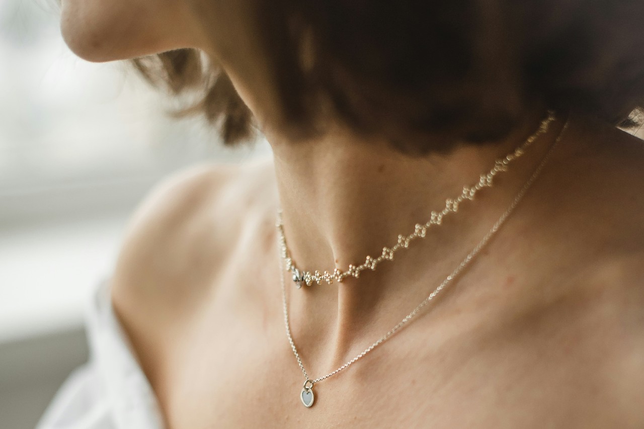 A close-up of a woman's neck adorned with a delicate pearl choker and a fine chain necklace featuring a small, heart-shaped pendant.