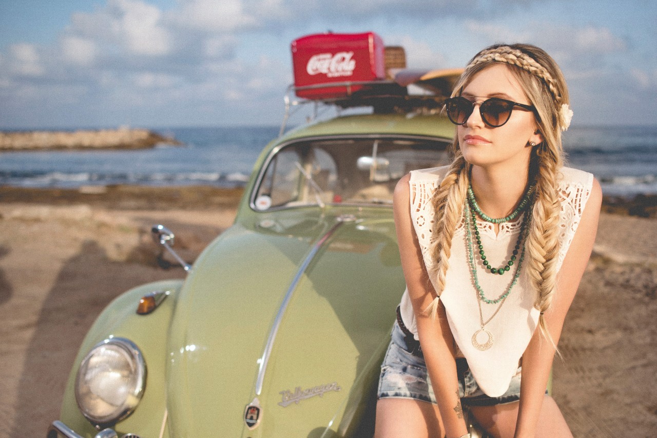 A boho-styled woman leans on a light green Volkswagon Beetle on the beach.