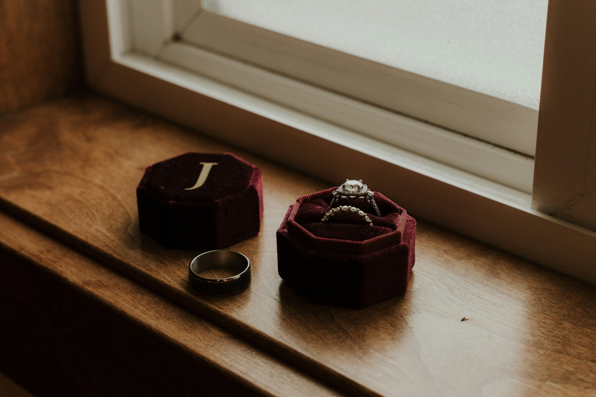 A velvet ring box sitting on a windowsill containing an engagement ring and matching band, and a men’s wedding band next to the box.